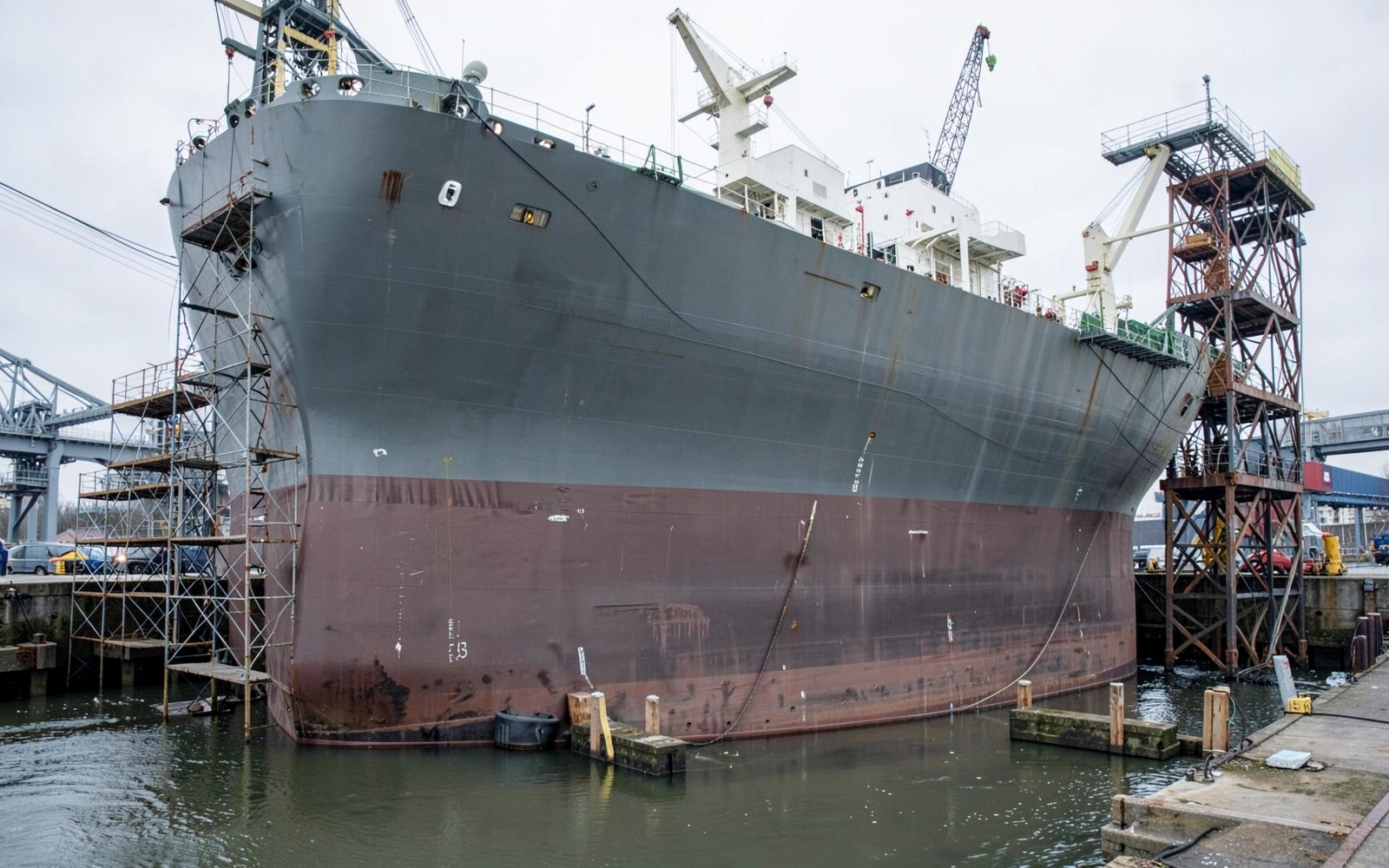 Large commercial ship hull in drydock with scaffolding and freshly coated underwater area. Illustrative industrial photograph.